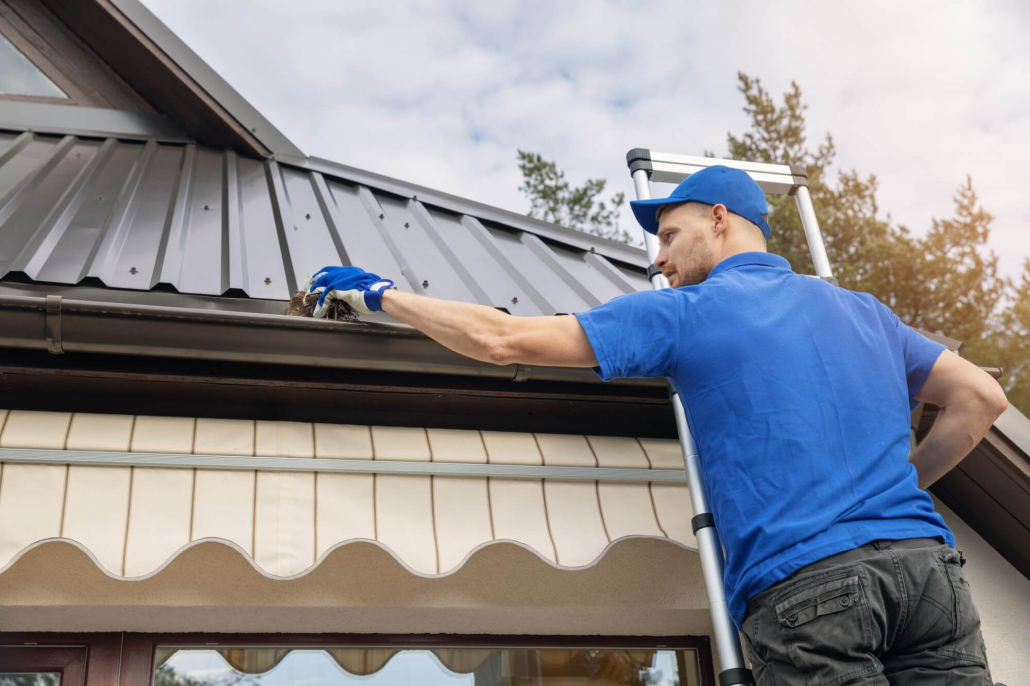 Gutter-cleaning-services A man standing on ladder and cleaning roof rain gutter from dirt.